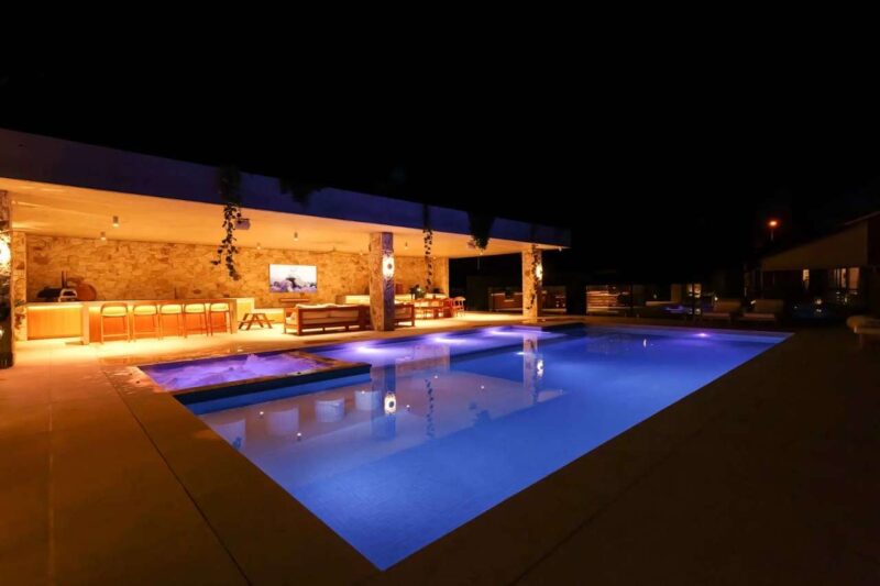 Luxury concrete swimming pool with glowing LED lights beside a modern outdoor entertaining area with bar, lounge, and TV setup at night.