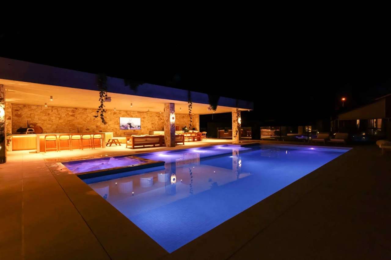 Luxury concrete swimming pool with glowing LED lights beside a modern outdoor entertaining area with bar, lounge, and TV setup at night.