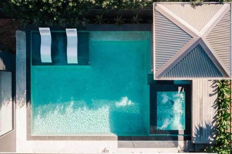 Aerial view of a modern geometric concrete swimming pool with built-in loungers, integrated spa, timber deck, and shaded pavilion in a Gold Coast backyard.